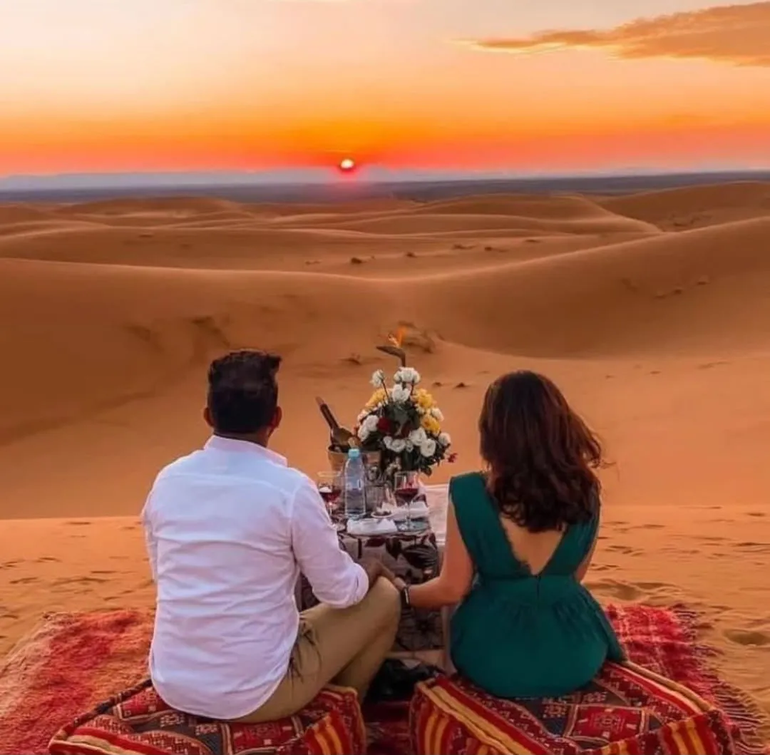 Coppia seduta sui tappeti davanti a un tavolino con fiori e calici, in cima a una duna di Merzouga al tramonto