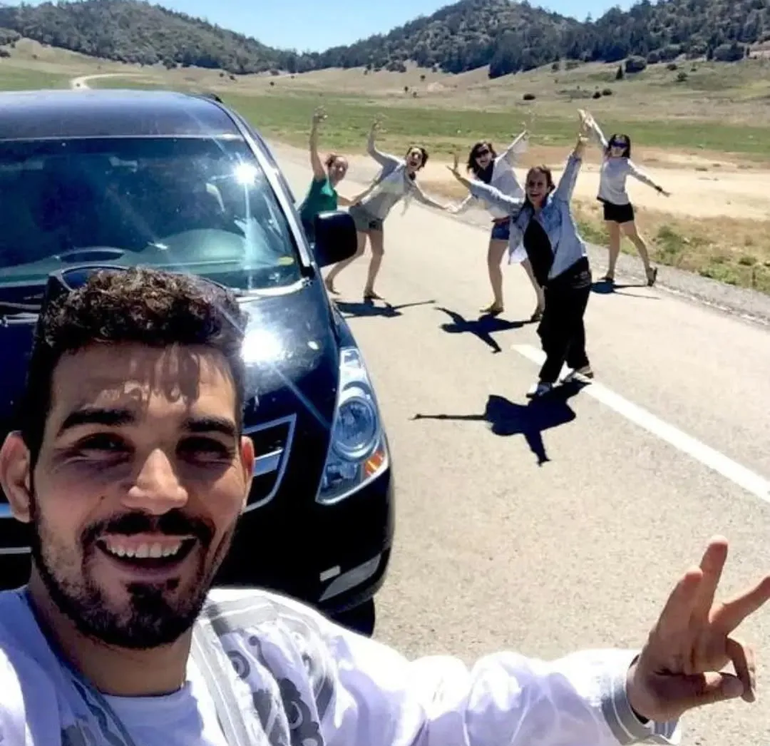 Autista del transfer privato accanto al furgone, con cinque viaggiatrici che salutano su una strada di montagna in Marocco