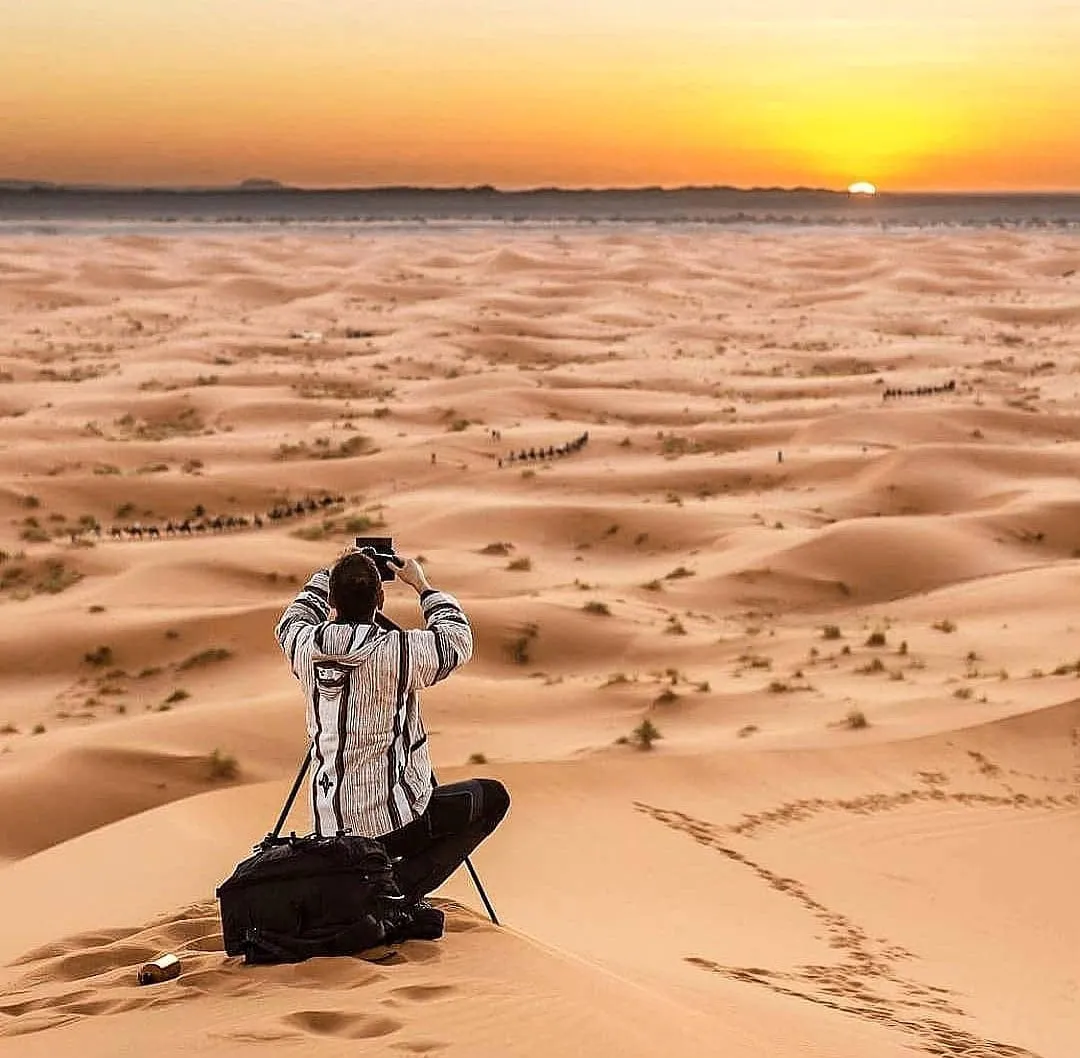 Fotografo con treppiede in cima a una duna di Merzouga, con carovane di dromedari in lontananza e il sole all’orizzonte