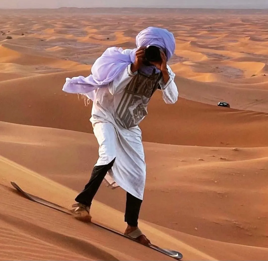 Uomo con turbante e djellaba che scende in sandboard da una duna di Merzouga mentre scatta una foto