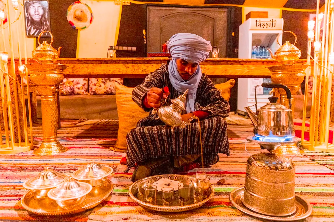 Uomo con il turbante che versa il tè alla menta dall’alto, tra teiere di ottone, nella tenda ristorante