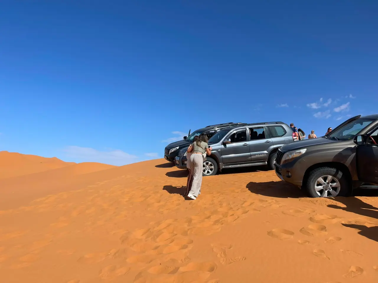 Several 4x4s parked on top of a dune, with guests walking down onto the sand