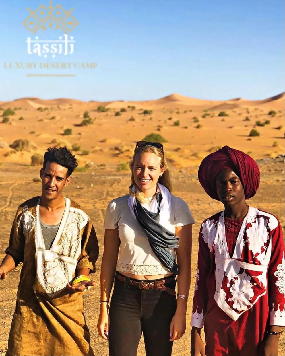 Una viaggiatrice sorridente tra due giovani berberi in abiti tradizionali, con le dune di Merzouga alle spalle