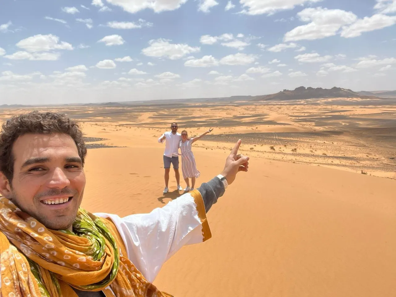 Un viaggiatore si scatta un selfie in cima a una duna di Merzouga, con una coppia alle spalle, in una giornata di sole